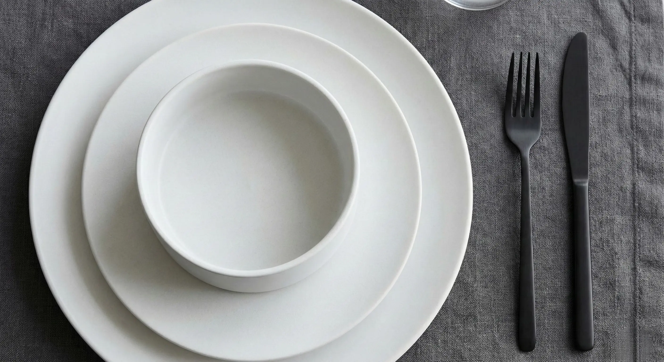 A close-up photograph of a modern, minimalist place setting featuring matte white porcelain dinnerware with clean straight sides